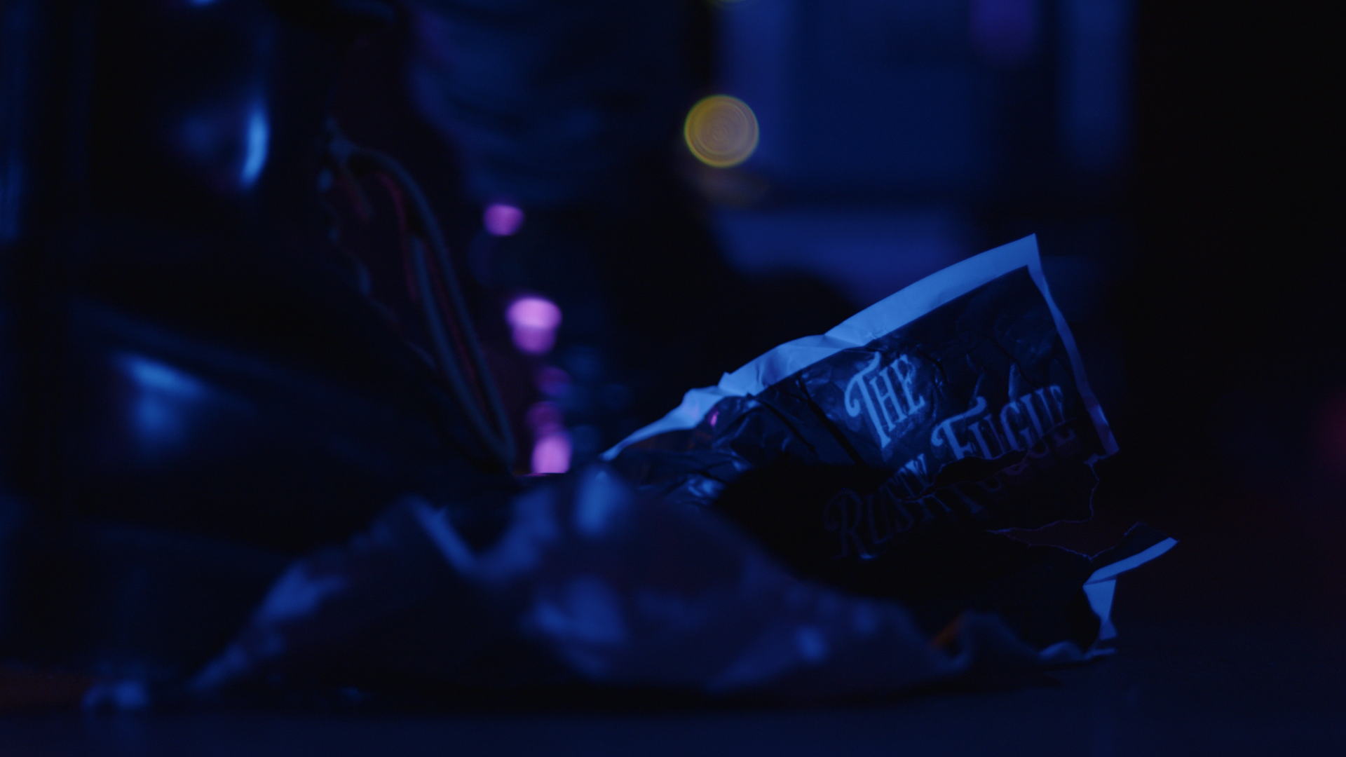 On the floor of a darkly-lit music venue, a boot tramples on a crumpled poster. The full details of the poster cannot be seen, but the name of the venue, "The Rusty Fugue" is visible in the top right corner, which sticks up