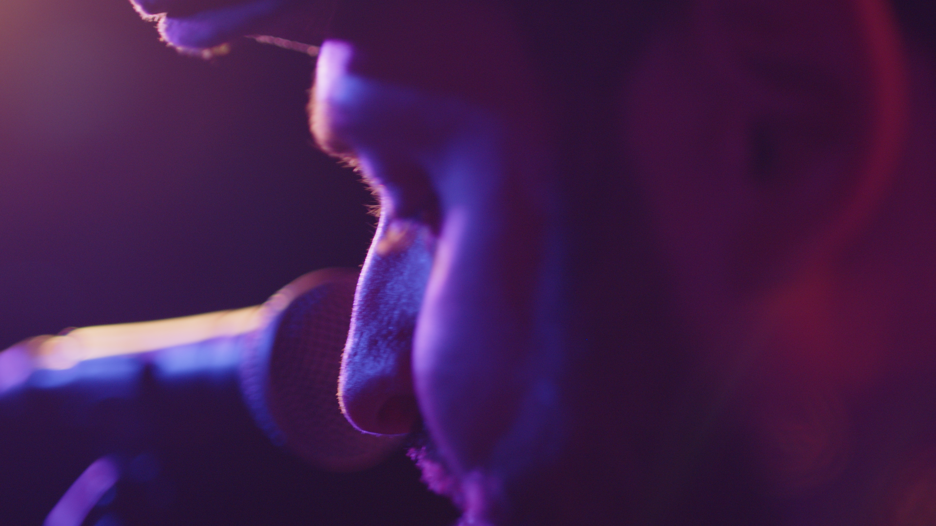 Close-up side profile of a musician as he sits on stage. The microphone is positioned close to his face and he is lit by warm, purple / orange stage lighting.