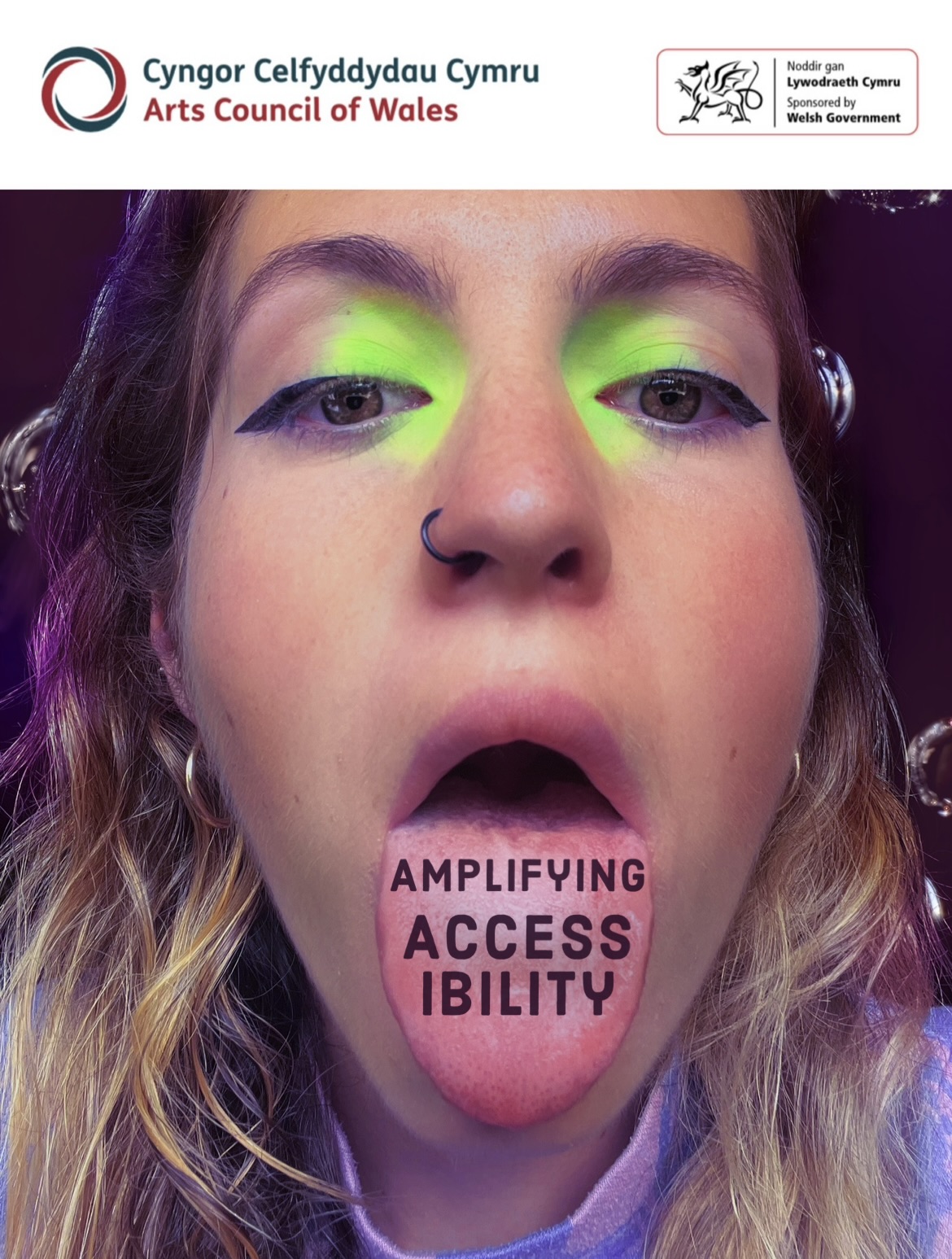 Face of a white female, with green eyeshadow and her tongue out. Her hair is light brown and the neck of her top is pink and purple. On her tongue it reads "Amplifying Accessibility" in black letters.