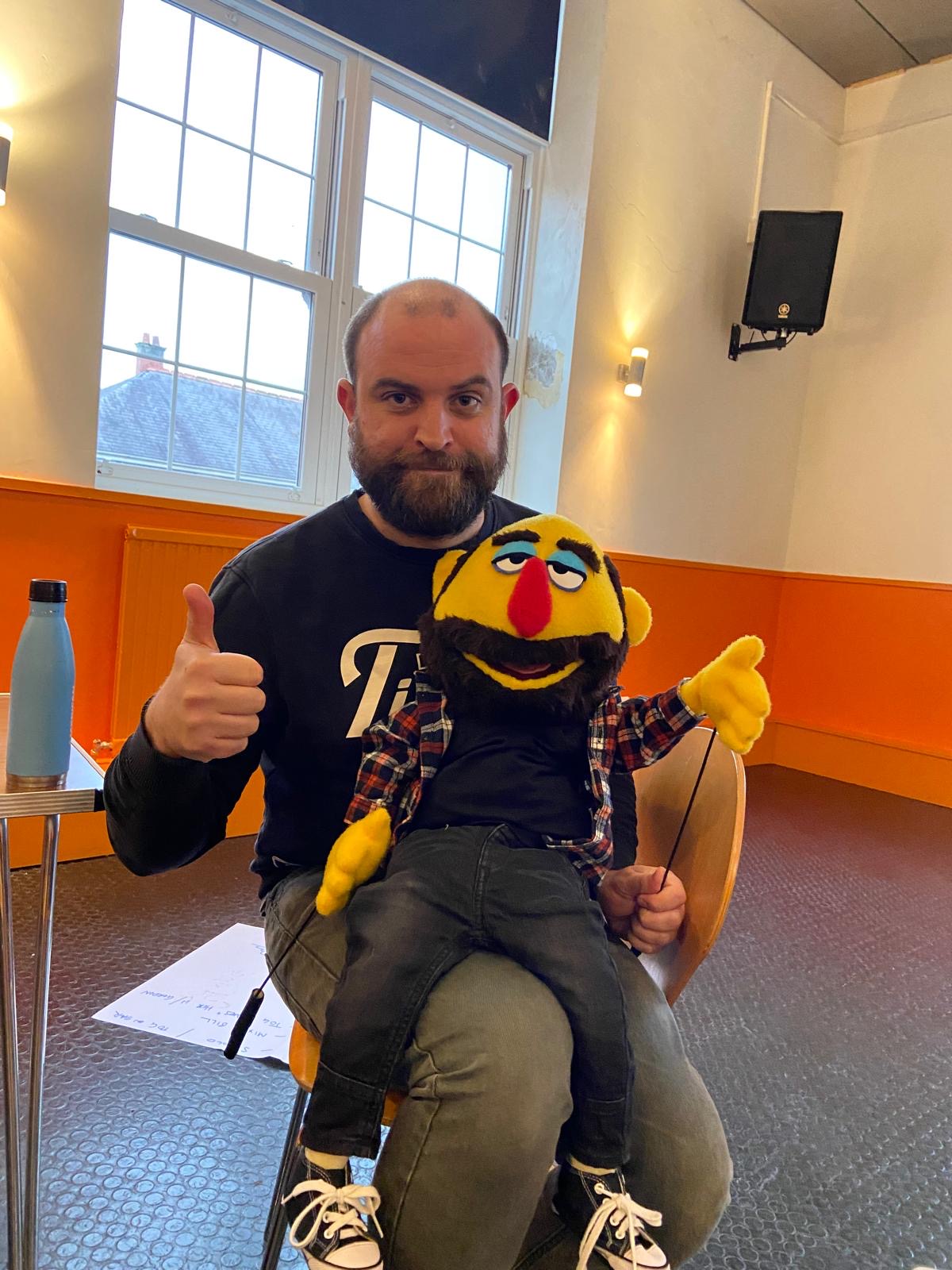 A man holds a yellow puppet version of himself on his lap