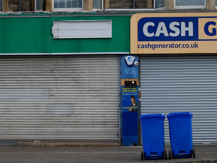 Two storefronts with closed metal shutters and two blue bins